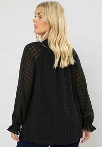 Schwarze Bluse mit langen, strukturierten, durchsichtigen Ärmeln. Verfügt über einen gerafften Ausschnitt und elastische Bündchen. Mit einem subtilen Rautenmuster gestaltet.