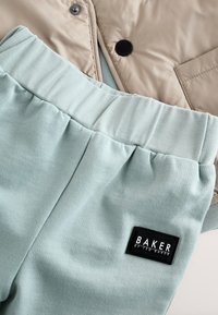 Veste beige clair à finition lisse portée au-dessus d'un pantalon de survêtement vert menthe doux, doté d'une étiquette de marque noire, d'une taille élastique et d'un design simple.