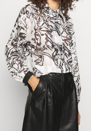 Blouse blanche transparente avec un motif abstrait noir, dotée d'un col et de manches longues, associée à un pantalon en cuir noir ajusté.