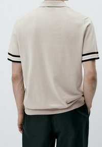 T-shirt polo beige en maille, avec des accents de rayures noires sur les manches et un col classique. Texture lisse, design décontracté.