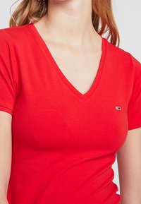 Rotes Baumwoll-T-Shirt mit V-Ausschnitt und kurzen Ärmeln, mit einem kleinen Logo auf der linken Brust. Glatte Textur und taillierte Passform.