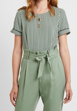 Vrouw draagt een korte mouwen blouse met patroon, ingestopt in salie groene broek met hoge taille en gebonden riem, en met gelaagde gouden kettingen.