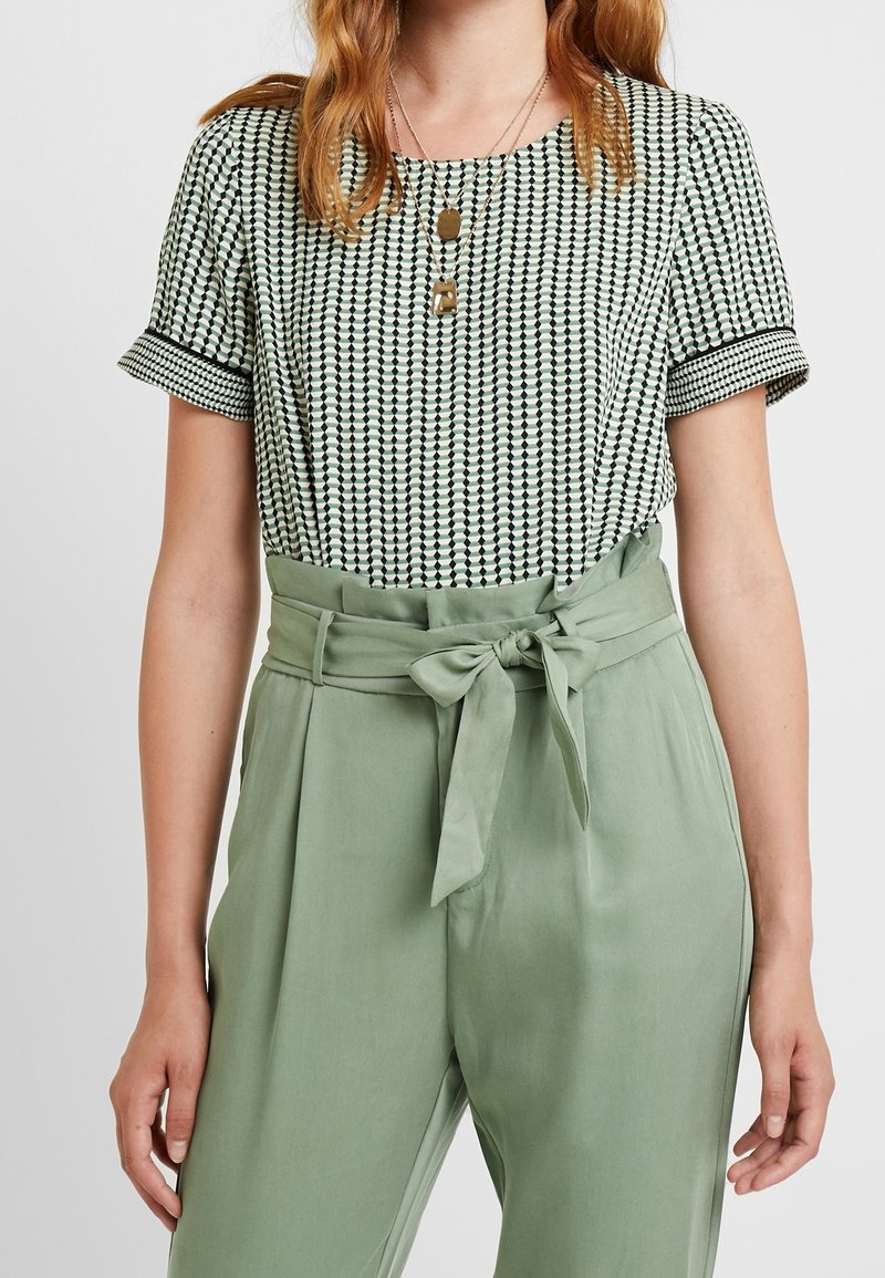 Femme portant un chemisier à manches courtes à motifs, rentré dans un pantalon vert sauge taille haute avec une ceinture nouée, et des colliers dorés superposés.