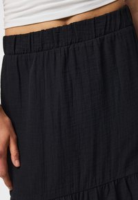 Gros plan d'une personne portant une jupe noire texturée avec une taille élastique et un top court blanc, montrant une partie de la main et de la taille.