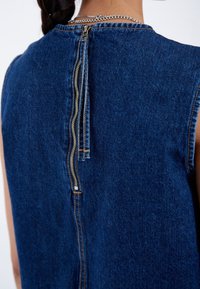 Prenda de mezclilla con un cuello alto, mangas cortas y un cierre posterior destacado en plata. Tejido de color azul oscuro con una textura suave.