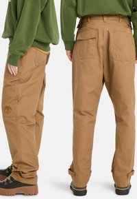 Tan cargo pants with a relaxed fit, featuring multiple pockets, side buttons, and a textured fabric. Paired with a green sweatshirt.