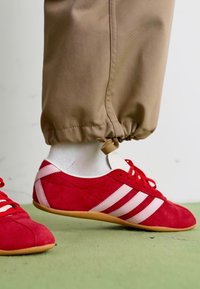 Červené semišové sportovní boty s bílými pruhy, s zaoblenou špičkou a texturovaným povrchem, spárované s béžovými kalhotami se strečovými manžetami.
