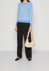 Kobieta ubrana w niebieską i białą w paski bluzkę z długim rękawem, czarne szerokie spodnie, czarne baleriny, trzymająca torebkę w kolorze kremowym z węzłowymi uchwytami.