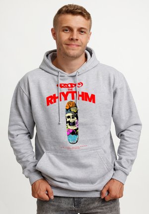 Jonge man draagt een lichtgrijze hoodie met rode "RHYTHM" tekst en een kleurrijk schedelontwerp op een skateboardgrafiek aan de voorkant.