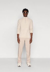 Muž oblečený v béžovém čtvrtzipovém fleece, béžových kalhotách, bílých teniskách a béžových slunečních brýlích stojí před čistým bílým pozadím.