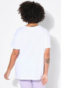 Hvid bomulds T-shirt med korte ærmer, rund halsudskæring og afslappet pasform. Viser et tatovering på ryggen nær halsen. Glat tekstur.