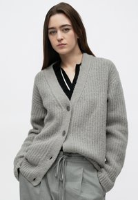 Šedý pletený kardigan s výstřihem do V, zapínáním na knoflíky a strukturovanou látkou. Nošený přes černý top, kombinovaný se šedými kalhotami na stažení.