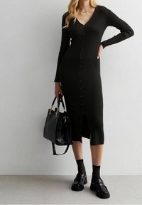 Robe en tricot côtelé noire avec col en V, manches longues, fermeture à boutons sur le devant et fente. Associée à un sac à main texturé noir et des chaussures à plateformes.