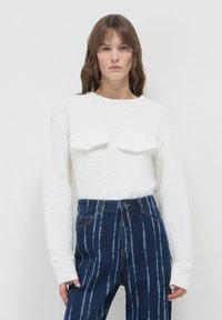 Maglione bianco strutturato con maniche lunghe e due tasche sul petto, abbinato a jeans blu a vita alta con strisce bianche verticali.