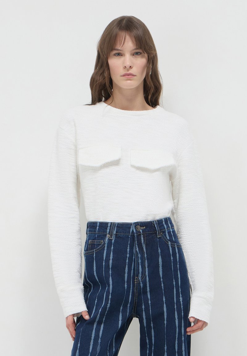 Maglione bianco strutturato con maniche lunghe e due tasche sul petto, abbinato a jeans blu a vita alta con strisce bianche verticali.