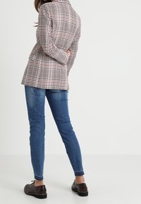 Blazer à carreaux beige et marron avec une coupe ajustée, associé à un jean bleu et des chaussures marron foncé. Poches visibles et épaules structurées.