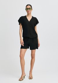 Top nero a maniche corte, scollo a V con maniche arricciate abbinato a pantaloni shorts neri. La modella indossa occhiali da sole verdi e tacchi verde oliva.