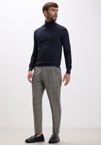 Ein Mann trägt einen strukturierten navyblauen Rollkragenpullover, graue karierte Hosen und schwarze gefranste Lederloafers, steht und schaut zur Seite.