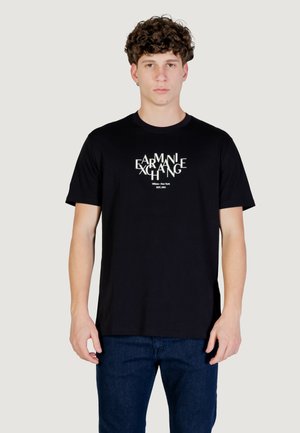 Jeune homme aux cheveux bouclés portant un t-shirt noir Armani Exchange avec un logo blanc, debout devant un fond gris clair uni.