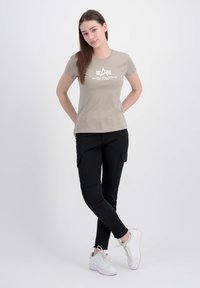 T-shirt beige à manches courtes avec un graphisme blanc "Alpha Industries" sur le devant, associé à un pantalon cargo noir et des baskets blanches.