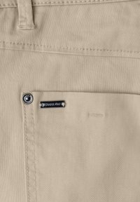 Poche en tissu beige avec un rivet en métal et une petite étiquette métallique rectangulaire gravée "Street One" sur un vêtement.
