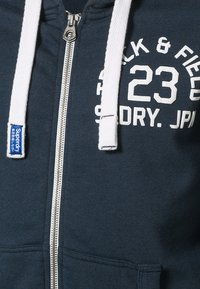 Sudadera azul marino con cremallera, con cordones blancos, bolsillo frontal y texto blanco que dice "TRACK & FIELD 23 SUPERDRY. JPN" en el pecho.