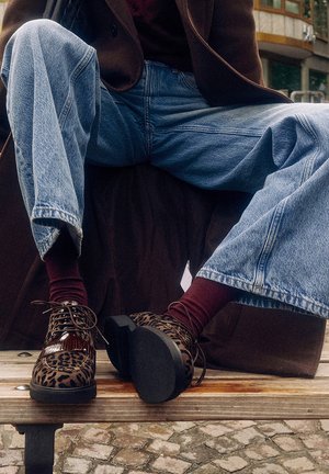 Chaussures à lacets à motif léopard avec des accents bruns brillants, une semelle texturée, portées avec un jean taille haute bleu et des chaussettes bordeaux, sur un banc en bois.