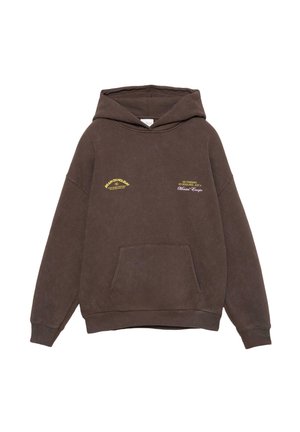 Personne portant un sweat à capuche marron, un pantalon blanc à jambes larges, des chaussures de randonnée beiges et des bagues à la main gauche sur un fond blanc.