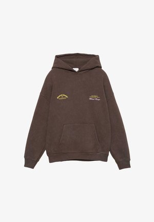 Personne portant un sweat à capuche marron, un pantalon blanc à jambes larges, des chaussures de randonnée beiges et des bagues à la main gauche sur un fond blanc.