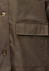 Tissu marron avec une grande poche rectangulaire et un rabat sur le côté droit, à côté de deux boutons cousus sur le côté gauche.