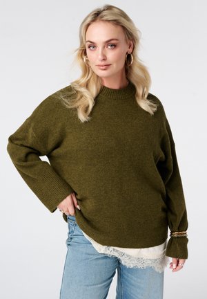 Blond vrouw met gouden oorringen, een oversized olijfgroene trui, een wit topje met kant en lichtblauwe spijkerbroek tegen een lichte achtergrond.