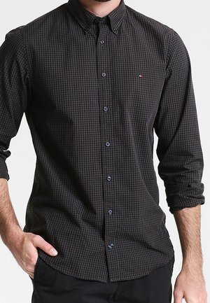 Mann trägt ein langärmliges, dunkel kariertes Hemd mit hochgekrempelten Ärmeln und einer Hand in der Tasche.