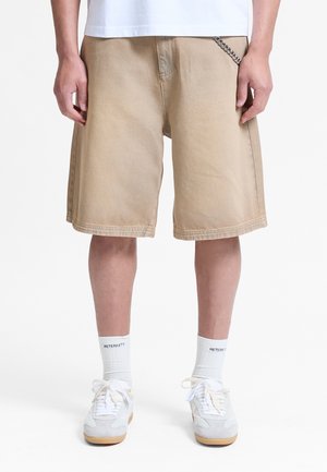 Persona che indossa pantaloncini beige fino al ginocchio, calzini bianchi con scritta, scarpe da ginnastica bianche e una camicia bianca, in piedi davanti a uno sfondo uniforme.