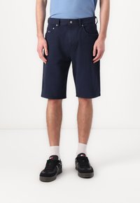 Mørkeblå bomullsshorts med rett snitt, med frontlommer og knappestenging. Brukt med svarte sneakers og hvite sokker.