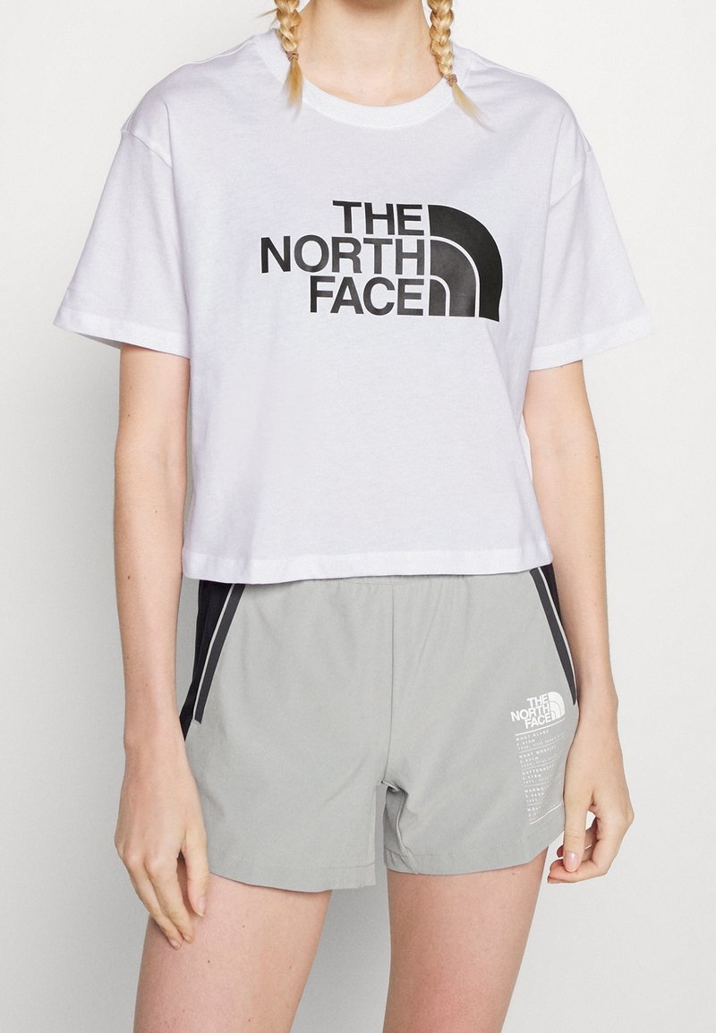 Bílý zkrácený tričko s černým logem "THE NORTH FACE", spárované se šedými šortkami s bočními černými akcenty a malým logem na přední straně.