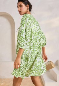 Rochie verde cu un model abstract alb, dintr-un material texturat, cu mâneci lungi și o croială lejeră, asortată cu o mică geantă țesută.