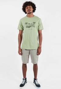 T-shirt en coton vert clair avec un motif de palmier et le texte "Oxbow Permanent Vacation", porté avec un short cargo beige et des baskets noires.