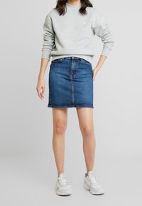 Sweat-shirt gris clair à col rond avec logo brodé, associé à une jupe en jean bleu foncé. Des baskets blanches complètent la tenue.