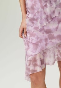 Lila Batik-Kleid mit gestuften Rüschen, aus durchscheinendem Stoff gefertigt. Weiche Textur; Akzente am Saum und entlang der Kanten erzeugen Bewegung.