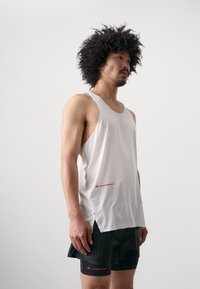 White athletic tank top with dotted texture, featuring a round neckline and side slits. Red logo accent on the lower front. Black shorts with red details.