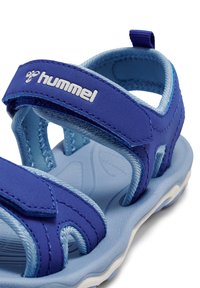 Niebieska sandał z regulowanymi paskami i wyściełaną wkładką. Wierzch wykonany z mieszanki gładkich i teksturowanych materiałów. Logo widoczne na pierwszym planie.
