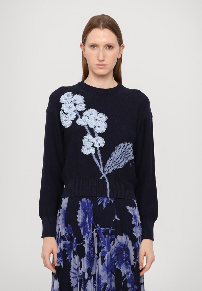 Navy sweater with light blue floral embroidery, cropped design, ribbed cuffs, and hem. Model paired with a floral pleated skirt.