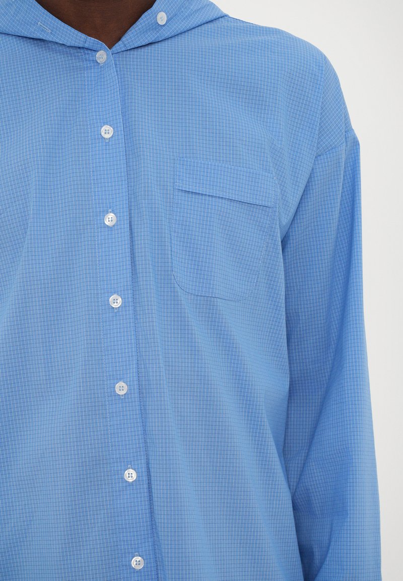 Camisa abotonada de color azul claro con capucha, patrón a cuadros y un solo bolsillo en el pecho. Fabricada en tela suave con un corte relajado.