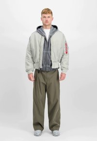 Młody mężczyzna stojący, ubrany w jasnoszary bomber jacket na szary sposób dzianinowy z kapturem, oliwkowe szerokie spodnie i szare sneakersy na jednolitym tle.
