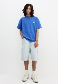 Blå bomulds t-shirt med et lille logo, parret med lyse denimshorts og hvide sneakers. Modellen står med hænderne i lommerne foran en enkel baggrund.