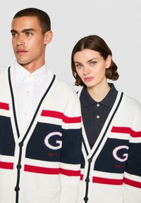 Hvit kardigan med marineblå og røde striper, med en stor "G" logo. Båret over en hvit skjorte med knapper og en marineblå polo, med knapper og lommer synlige.