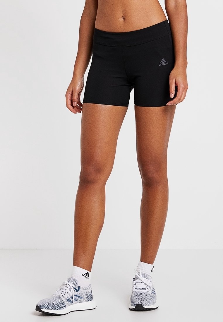 Pantalones cortos deportivos negros con un diseño ajustado, una cinturilla ancha y un pequeño logo gris en el lado izquierdo. Combinados con zapatillas grises y blancas.