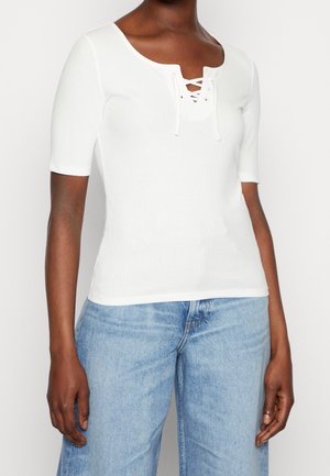 Kvinde iført en hvid ribbet kortærmet top med snørelukning ved halsudskæringen, kombineret med lyseblå denimjeans, stående med armene afslappet.