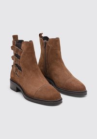 Botas de tobillo de ante marrón con puntera puntiaguda, cremallera lateral y tres correas decorativas en el lado exterior; suela de goma y detalles de costura.