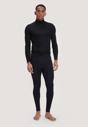 Mann steht barfuß, trägt einen enganliegenden schwarzen Langarm-Rollkragenpullover und schwarze Leggings vor einem einfarbigen hellen Hintergrund.